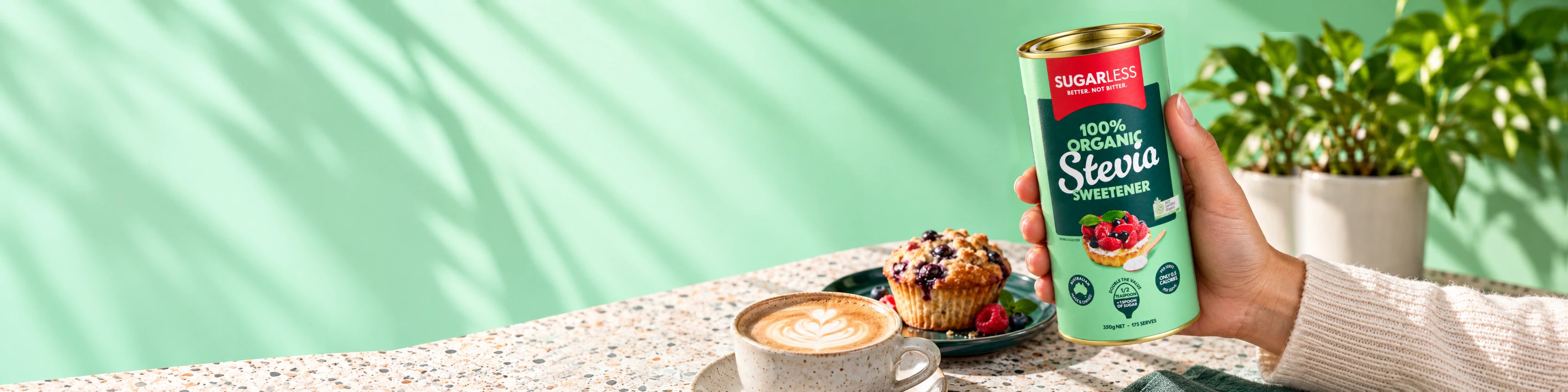 Hand holding Sugarless Organic Stevia beside a latte and blueberry muffin.